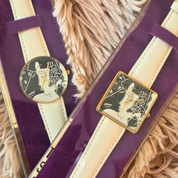 Two vintage-style wristwatches displayed on purple velvet stands against a beige faux-fur background. One watch has a round gold-tone case with a black dial and cream strap, and the other has a square gold-tone case with a black-and-white illustration and cream strap.